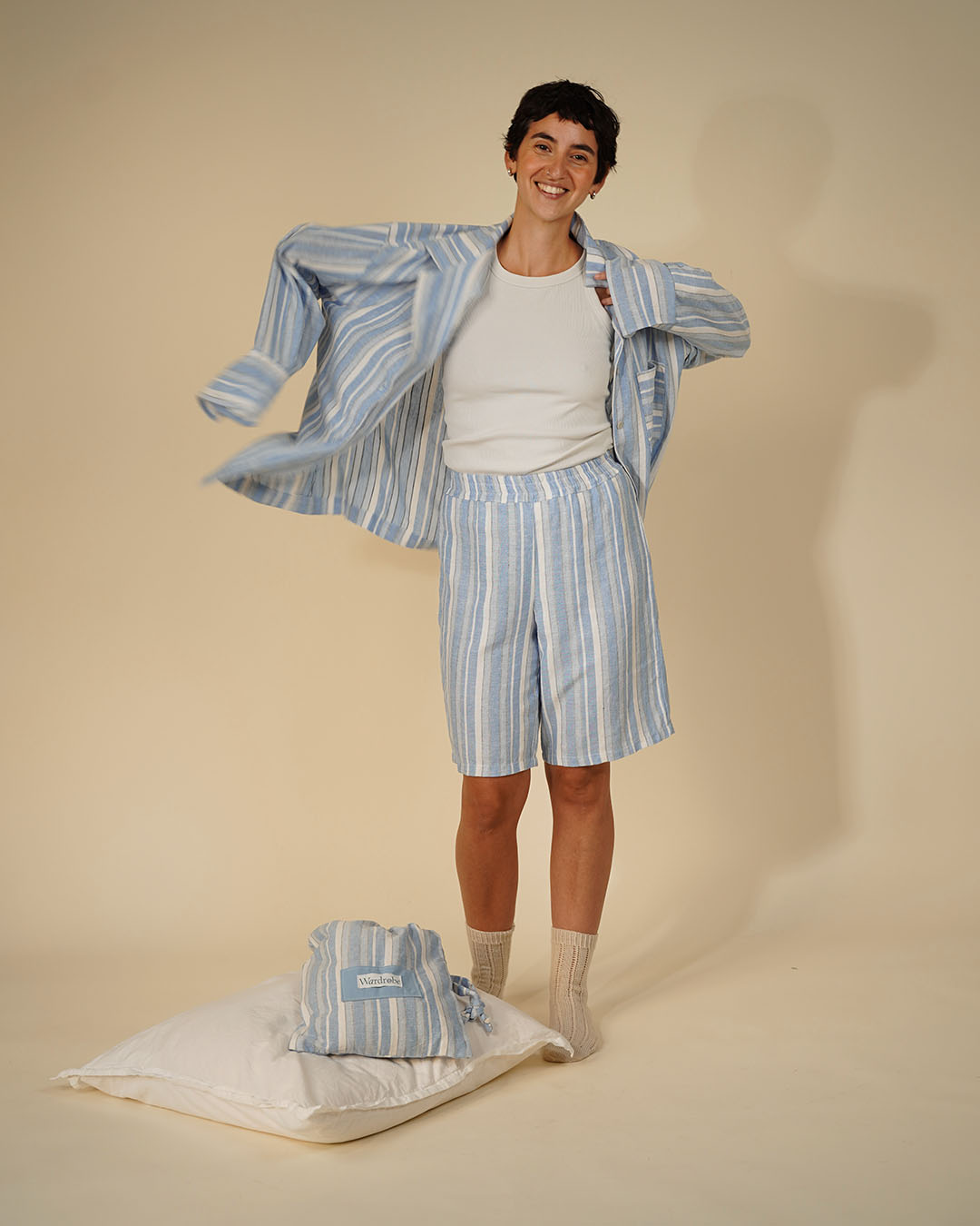 A smiling man models a light blue and white striped pajama set, including a long-sleeve shirt and shorts, over a white t-shirt. A matching folded pajama set rests on a white pillow on the floor next to him, set against a solid beige studio background.