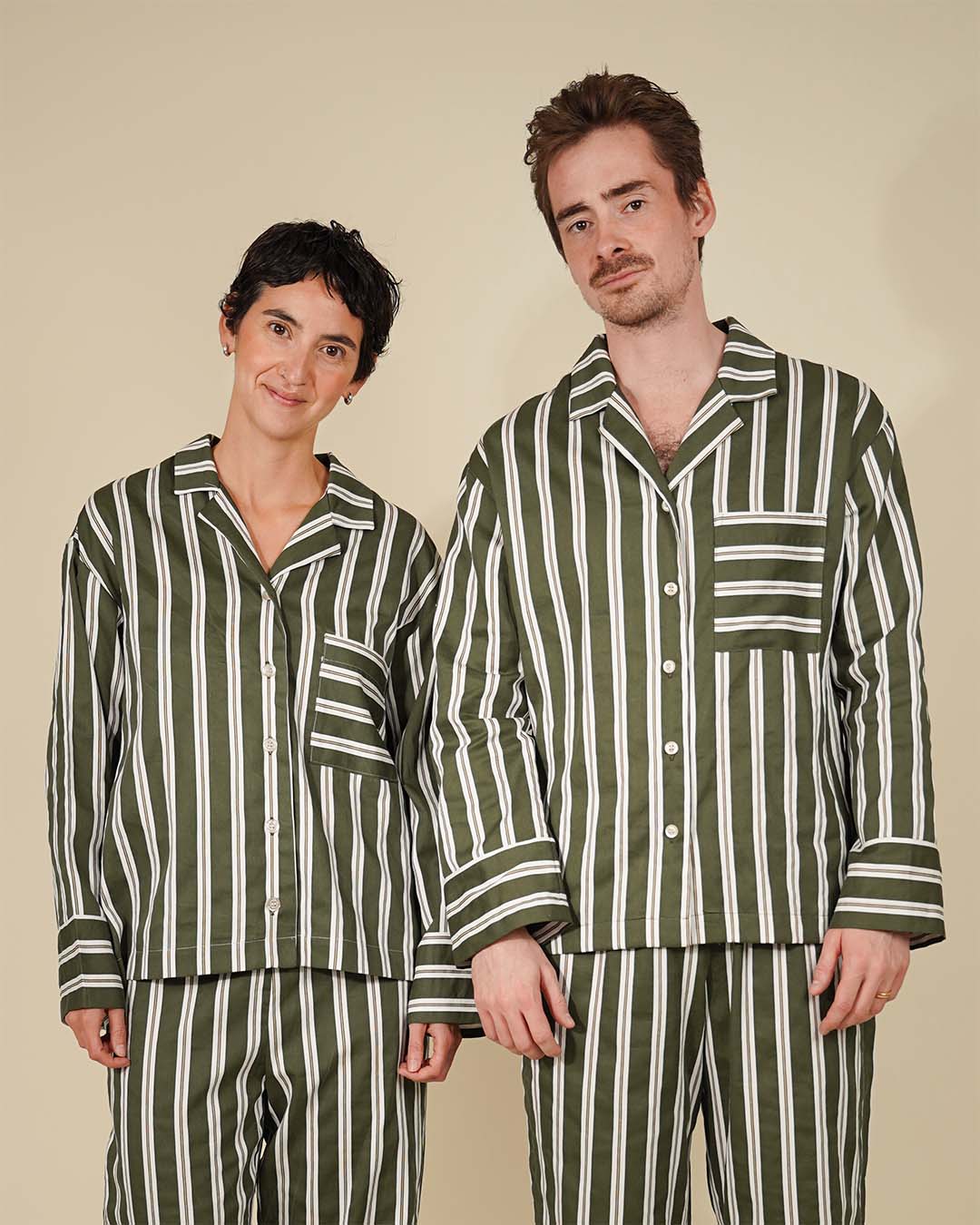 A man and a woman modeling matching green and white vertically striped pajama sets against a solid beige background.