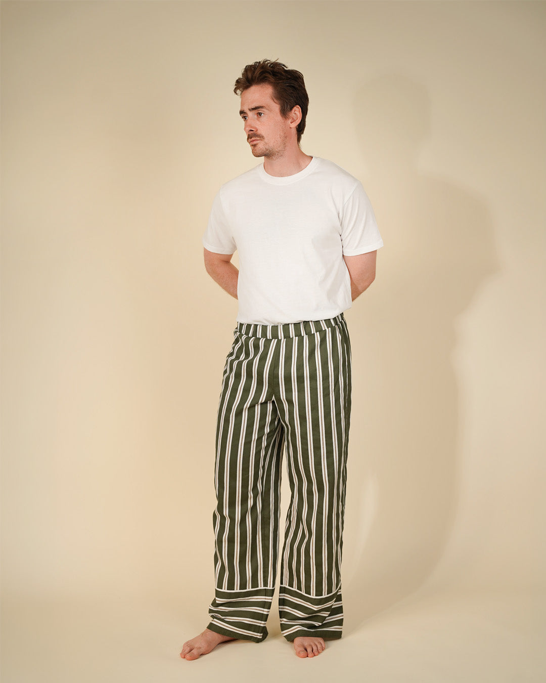 A man stands barefoot against a plain beige studio background, wearing a plain white short-sleeve t-shirt and loose vertical green-and-white striped pants.