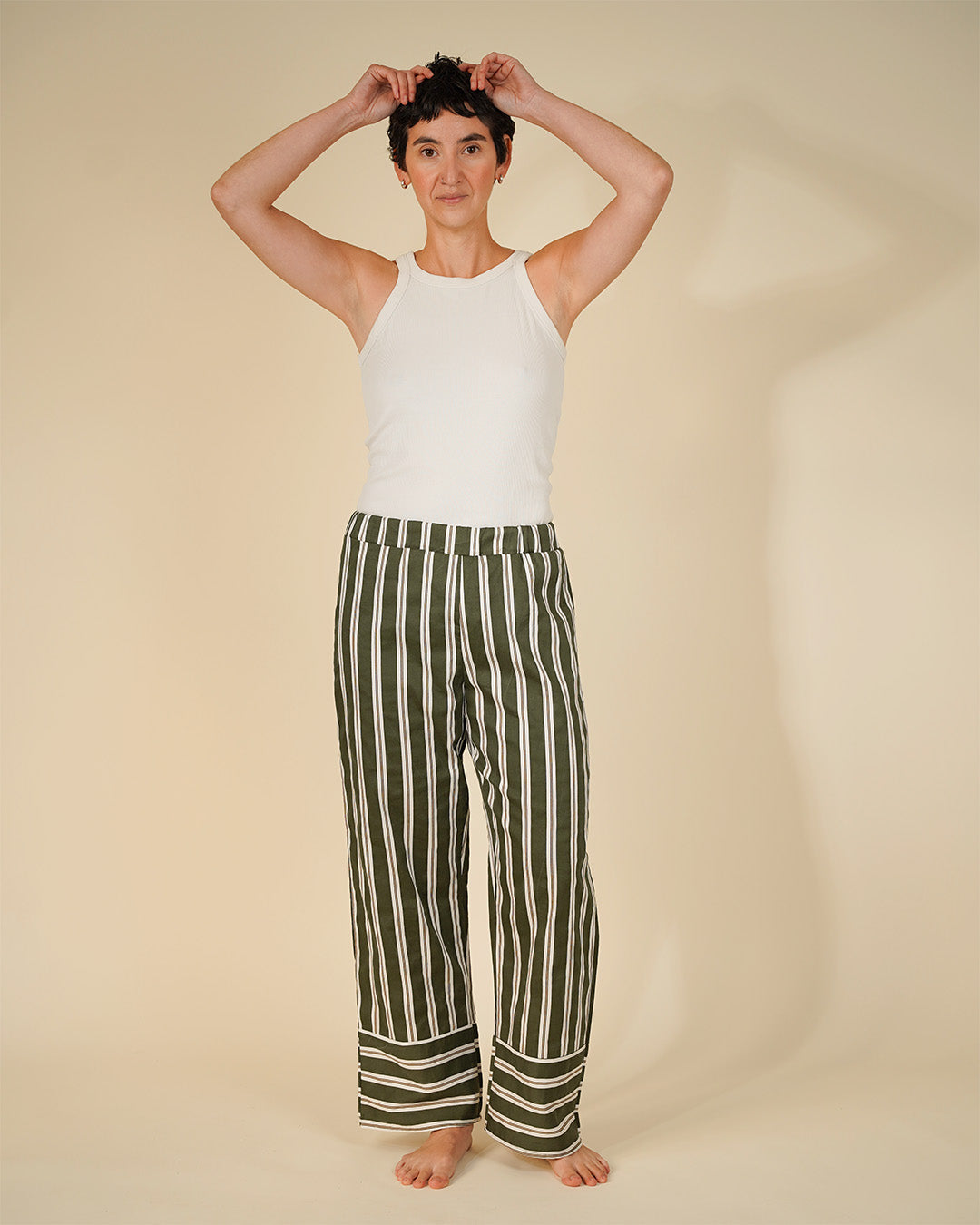 A model stands barefoot against a warm beige studio background, wearing a white sleeveless top and wide-leg trousers with vertical olive green and off-white stripes. The model has short dark hair and is holding it up with both hands.
