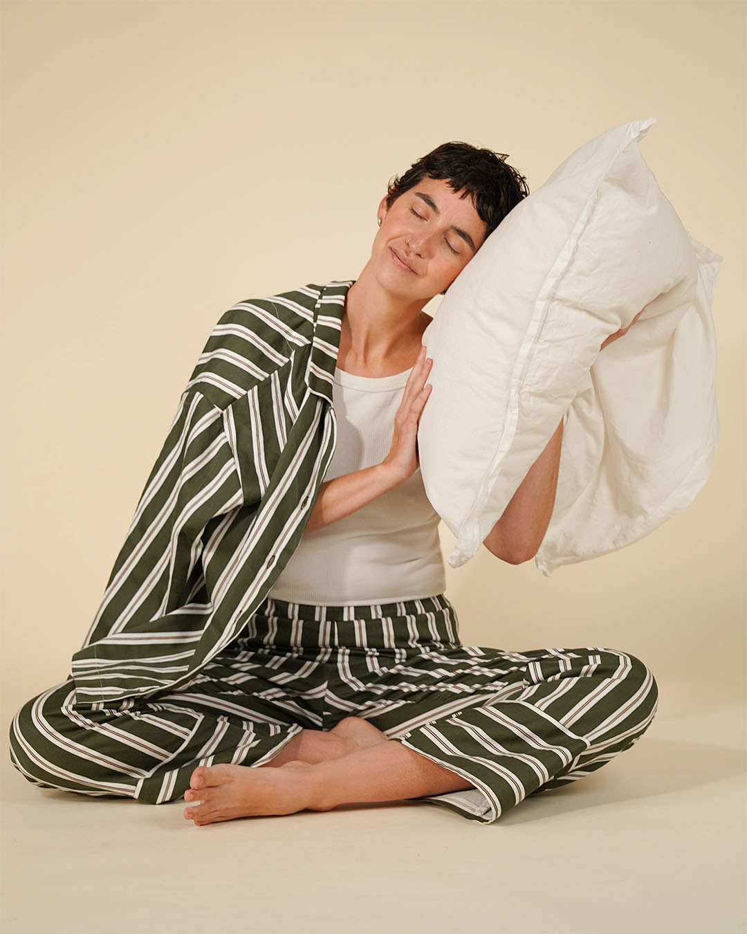 A woman sits cross-legged on a beige studio floor wearing a matching green and white striped pajama set, with a white pillow against her head.