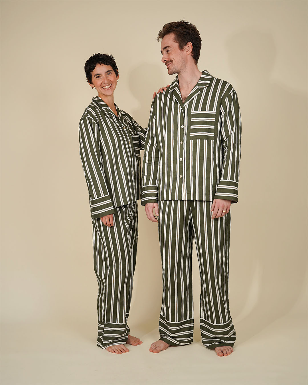 A man and a woman wearing matching striped pajama sets, standing barefoot against a plain beige studio background. The pajamas feature vertical stripes in dark green and off-white.